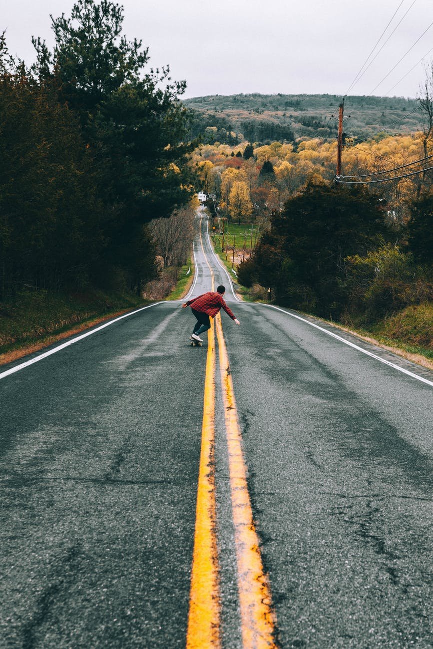 man in red shirt riding skateboard on empty country road